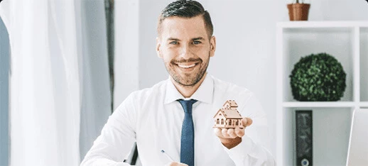 A well dressed guy is holding a small dummy of a house and smiling