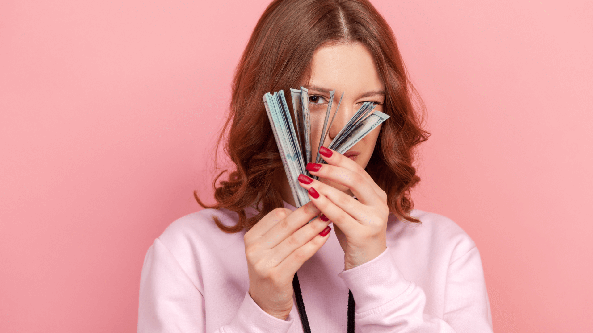 A girl in a single frame with pink background and pink hoodie is holding a bundle of cash and looking through the bundle cash closing her one eye